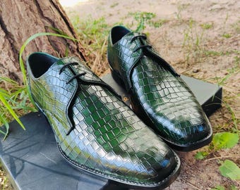 May include: A pair of green leather dress shoes with a textured, crocodile-like pattern. The shoes have black laces and a sleek, polished appearance. They are displayed on a dark surface, suggesting a formal or upscale style.