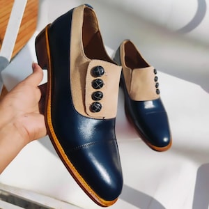 May include: Two-tone dress shoes in navy blue and tan. The shoes have a navy leather toe and vamp, a tan suede panel with four black buttons, and brown soles with a yellow border.