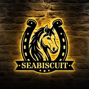 Pode incluir: Arte de parede em metal preto com a cabeça de um cavalo dentro de uma ferradura. A palavra "SEABISCUIT" é exibida abaixo, com três estrelas. A arte é iluminada por uma luz dourada quente.