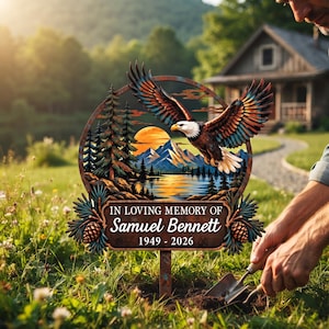May include: A circular metal memorial garden stake featuring a bald eagle in flight above a mountain lake landscape. The plaque includes the text IN LOVING MEMORY OF Samuel Bennett 1949 - 2026, framed by pine trees and pinecones.