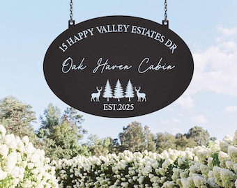 May include: Black oval metal sign with white text, hanging from chains. The sign reads "15 Happy Valley Estates Dr Oak Haven Cabin EST.2025" with deer and tree graphics. The sign is hanging in front of a path lined with white flowers.