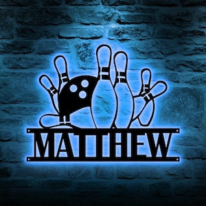 May include: Black metal bowling-themed wall art with a blue neon glow. The design features a bowling ball, pins, and a hand. The name "MATTHEW" is displayed below the bowling graphic. The art is mounted on a textured brick wall.