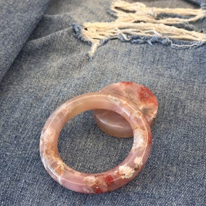 Puede incluir: Un brazalete translúcido rosa y blanco con inclusiones rojas y blancas. El brazalete es circular y descansa sobre un objeto circular más pequeño y a juego. El fondo es una tela vaquera azul con zonas desgastadas.