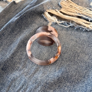 May include: A translucent agate bracelet with brown, orange, and white patterns. The circular bracelet sits on a denim surface. A matching agate stone is also visible.