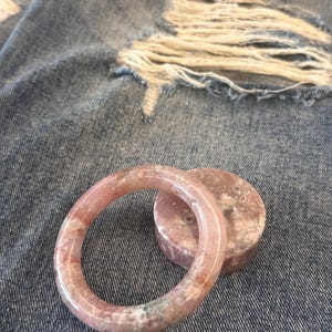 Puede incluir: Un brazalete de piedra preciosa rosa y blanca y una pieza redonda a juego. El brazalete es circular y tiene un aspecto translúcido. Las piezas de piedras preciosas están sobre un fondo de mezclilla.