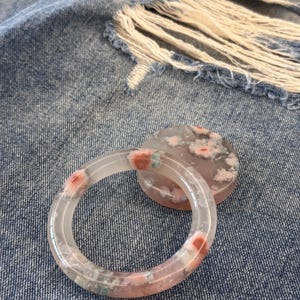 Puede incluir: Un brazalete transparente y un colgante redondo a juego, ambos con un estampado floral. El brazalete y el colgante presentan diseños florales rosas y azules. La joyería está sobre un fondo de mezclilla azul.