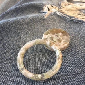 Può includere: Un braccialetto rotondo e decorato con un elemento di accento circolare abbinato. Il braccialetto e l'accento presentano un mix di colori bianco, verde e beige. Il gioiello è posto su uno sfondo in denim blu.