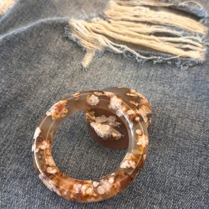 Puede incluir: Dos pulseras de ágata translúcida con un patrón floral en tonos marrón, blanco y dorado. Las pulseras son circulares y están sobre un fondo de mezclilla desgastada.