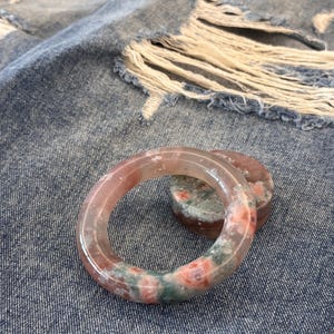 Puede incluir: Un brazalete de piedra rosa y verde translúcida y una piedra redonda a juego. El brazalete tiene un diseño circular con una mezcla de tonos rosas, verdes y blancos. La piedra descansa sobre una superficie de mezclilla azul.