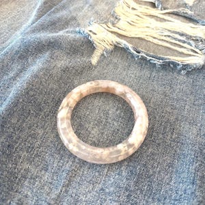 Puede incluir: Una pulsera redonda y transparente con un estampado floral en tonos blancos y rosas. La pulsera descansa sobre un fondo de mezclilla desgastada. La pulsera es un accesorio de moda.