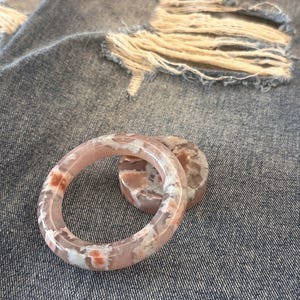 Puede incluir: Un par de pulseras de ágata rosa y blanca y una piedra redonda a juego. Las pulseras tienen un aspecto veteado con tonos de rosa, blanco y naranja. Los artículos se exhiben sobre un fondo de mezclilla desgastada.