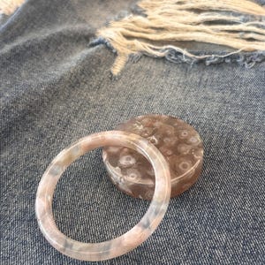 Puede incluir: Un brazalete de ágata rosa claro y blanco y un recipiente redondo a juego con un estampado floral. El brazalete es circular y descansa sobre una superficie de mezclilla desgastada.