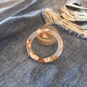 Könnte beinhalten: Ein transparenter Armreif aus Harz mit einem floralen Muster in Rosa- und Orangetönen. Der Armreif liegt auf einer Denim-Oberfläche mit Used-Details.