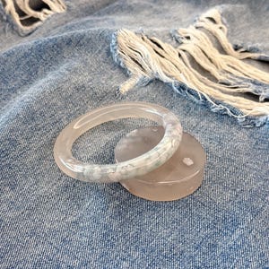 Könnte beinhalten: Ein durchscheinender Jade-Armreif mit hellgrüner Marmorierung liegt auf einem passenden, runden Jade-Untersetzer. Der Schmuck ist vor einem Hintergrund aus zerrissenem blauen Denim platziert.