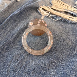 Può includere: Un braccialetto rotondo trasparente con sottili accenti floreali rosa e verdi. Il braccialetto è posto su uno sfondo di jeans blu strappati. È visibile anche una pietra con motivo floreale abbinato.