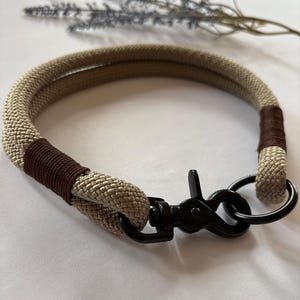 May include: A tan and brown rope dog collar with a black metal clasp and ring. The collar is made of a thick, woven rope material and has brown accents. The clasp and ring are black.