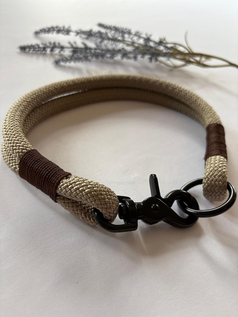 May include: A tan and brown rope dog collar with a black metal clasp and ring. The collar is made of a thick, woven rope material and has brown accents. The clasp and ring are black.
