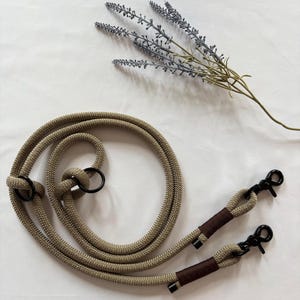 May include: Tan rope strap with black metal clasps and rings. The rope is knotted in several places. The clasps are attached to brown wrapped sections. A sprig of lavender is in the background.