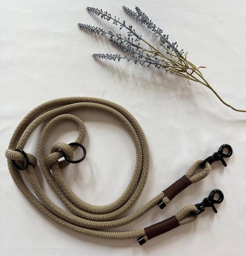 May include: Tan rope strap with black metal clasps and rings. The rope is knotted in several places. The clasps are attached to brown wrapped sections. A sprig of lavender is in the background.