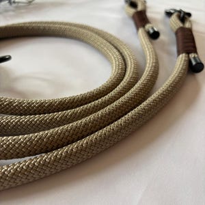 May include: Tan rope camera strap with brown leather accents and black metal hardware. The rope is thick and textured, and the strap has a loop at each end for attaching to a camera. The background is white.