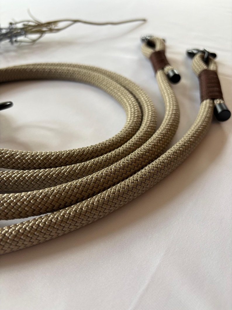 May include: Tan rope camera strap with brown leather accents and black metal hardware. The rope is thick and textured, and the strap has a loop at each end for attaching to a camera. The background is white.