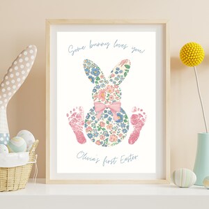 Puede incluir: Impresión enmarcada con un diseño floral de conejito, un lazo rosa y huellas de pies rosas. El texto en cursiva dice "Some bunny loves you" y "Olivia's first Easter". La impresión está en un marco de madera clara.