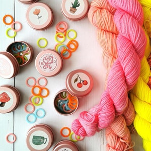 May include: Assortment of small pink metal tins with various floral and nature-themed designs. Each tin contains colorful plastic stitch markers. Two skeins of yarn, one pink and orange ombre and one yellow, are on a white wooden surface.