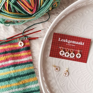 May include: A close-up of a set of red and white stitch markers with a gold cross design. The stitch markers are on a white card with the text "Leukegemaakt by Ingrid Peters" and a website address. The stitch markers are also on a white plate with a decorative pattern.