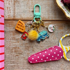 Op de afbeelding: Een metalen sleutelhanger met een lichtgroene karabijnhaak en diverse bedels, waaronder een oranje wafel, een rode thermoskan met bloemenmotief, een gele pompon, een blauwe camper en een ronde bedel met een bloemetje en een witte kraal.