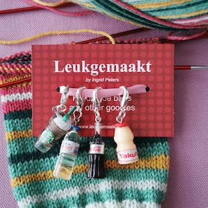 Op de afbeelding: Vier kleurrijke miniatuur drankcharms met zilveren ringen, bevestigd aan een roze-wit gestreepte gebreide sok. De charms zijn gevormd als een Starbucks Frappuccino, een fles Evian water, een Coca-Cola fles en een Yakult yoghurt drank. De charms zijn gelabeld met de merknamen. De kaart achter de charms staat "Leukegemaakt by Ingrid Peters".