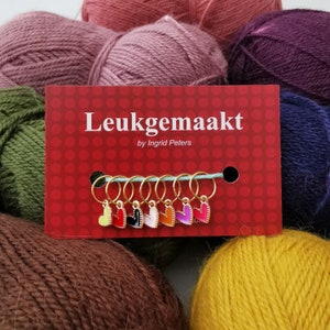 May include: A set of eight gold heart-shaped stitch markers with colorful enamel accents. The markers are displayed on a red card with the text "Leukegemaakt by Ingrid Peters".