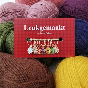 May include: A red card with the text "Leukgemaakt" and daisy stitch markers. The daisy stitch markers have gold rings and colorful enamel flowers in blue, red, purple, yellow, and green. The background features various skeins of yarn.