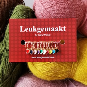May include: A set of 10 gold heart-shaped stitch markers with colorful enamel accents. The markers are displayed on a red card with the text "Leukegemaakt by Ingrid Peters" and the website address "www.leukegemaakt.com".