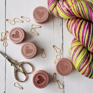 Op de afbeelding: Vijf kleine roze metalen containers met een hartvormig ontwerp en bloemmotieven. De containers staan op een wit houten oppervlak met gouden veiligheidsspelden en een bolletje veelkleurig garen.