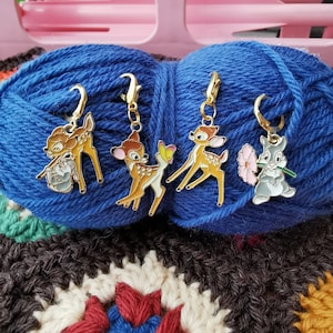 Pode incluir: Quatro pingentes de metal coloridos com personagens da Disney, incluindo Bambi, Tambor e um coelho, presos a anéis dourados. Os pingentes são exibidos em uma bola de lã azul.