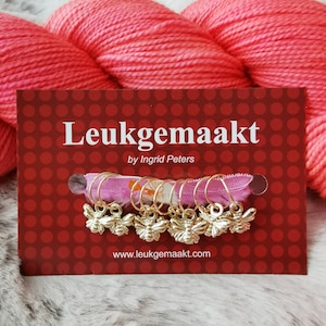 May include: A red ball of yarn with a card featuring the text "Leukegemaakt by Ingrid Peters" and a row of gold bee stitch markers on pink ribbon.