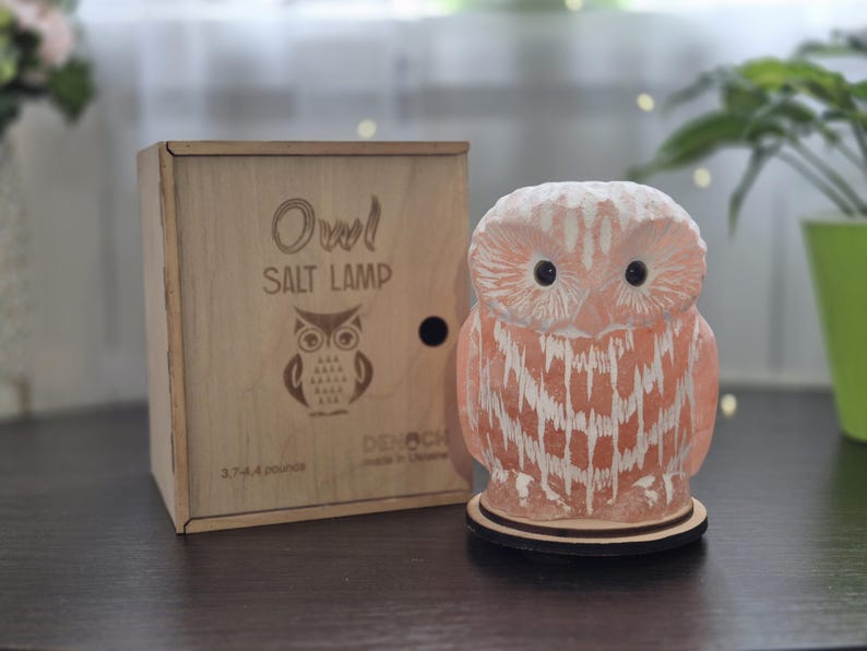 May include: A pink owl-shaped salt lamp with white accents, sitting on a wooden base. Beside it is a wooden box with the words "Owl Salt Lamp" and an owl illustration. The box also indicates the lamp weighs 3.7-4.4 ounces.