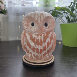 May include: A decorative owl figurine in shades of orange and white, standing on a wooden base. The owl has large, dark eyes and a textured surface. The figurine is a decorative item.
