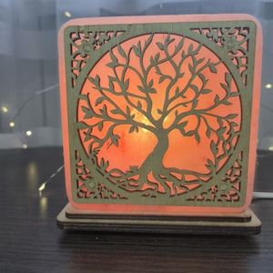 May include: A square, wooden lamp with a tree of life design. The tree is cut out of the wood, and the light shines through the design. The lamp has a warm, orange glow and sits on a wooden base.