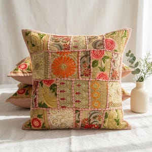 May include: A square throw pillow featuring a patchwork design of embroidered fabric panels in shades of gold, orange, pink, and green. The surface displays intricate floral and paisley patterns with raised pink stitching outlining each rectangular section.