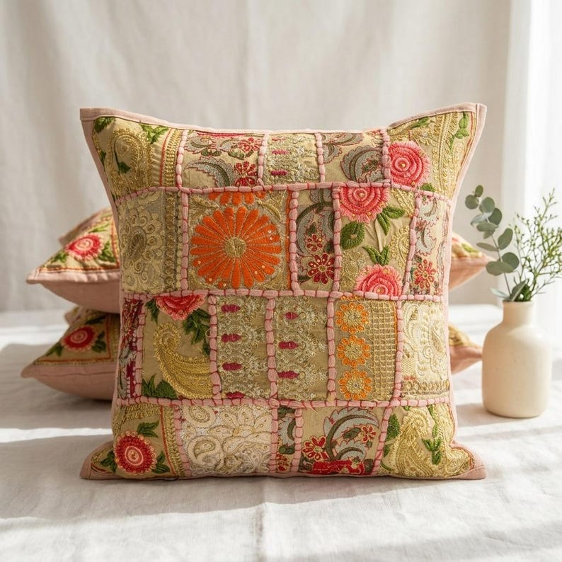 May include: A square throw pillow featuring a patchwork design of embroidered fabric panels in shades of gold, orange, pink, and green. The surface displays intricate floral and paisley patterns with raised pink stitching outlining each rectangular section.