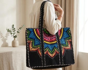 May include: A black tote bag featuring a vibrant, multi-colored mandala pattern in shades of blue, pink, green, and yellow. The bag has a textured surface, two long shoulder straps with a black and white checkered trim, and a rectangular shape.