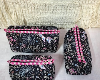 May include: Three black cosmetic bags with a floral and butterfly pattern. The bags have pink striped zippers and are in different sizes. The background is a cream-colored woven textile.