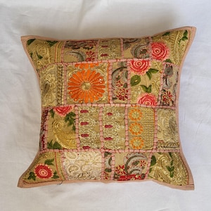 May include: A square decorative pillow cover featuring a patchwork design of gold-toned fabric panels. The surface is adorned with intricate embroidery, floral motifs in orange and pink, and metallic thread accents, all joined by pink stitching.