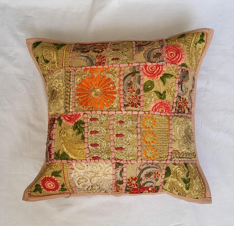 May include: A square decorative pillow cover featuring a patchwork design of gold-toned fabric panels. The surface is adorned with intricate embroidery, floral motifs in orange and pink, and metallic thread accents, all joined by pink stitching.