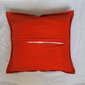 May include: A square throw pillow cover in a solid, vibrant orange color. The cover features a central horizontal opening with an overlapping fabric flap design, revealing a glimpse of the white inner pillow insert through the middle gap.