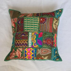 May include: A square throw pillow cover featuring a patchwork design of various fabric panels in shades of green, red, purple, and gold. The surface is decorated with intricate embroidery, small mirrors, and floral patterns stitched together.