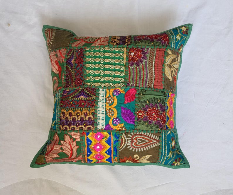 May include: A square throw pillow cover featuring a patchwork design of various fabric panels in shades of green, red, purple, and gold. The surface is decorated with intricate embroidery, small mirrors, and floral patterns stitched together.