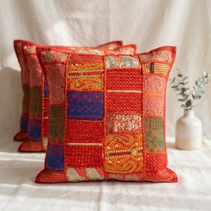 May include: Two square throw pillows featuring a patchwork design of various red, orange, blue, and green fabric panels. The surface is decorated with intricate embroidery, metallic thread stitching, and small reflective sequins in a grid pattern.