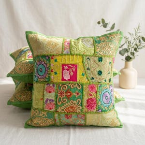 May include: A square throw pillow featuring a patchwork design of various green, yellow, and gold fabric panels. The surface is decorated with intricate embroidery, floral motifs, and small sequins in shades of pink, blue, and white.